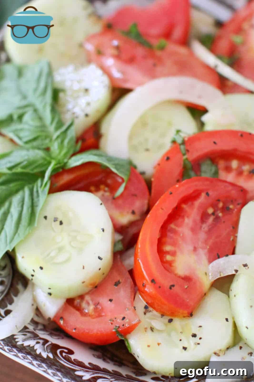 Closeup photo of marinated sliced tomatoes, cucumbers, and red onion in a bowl, garnished with a fresh basil leaf.