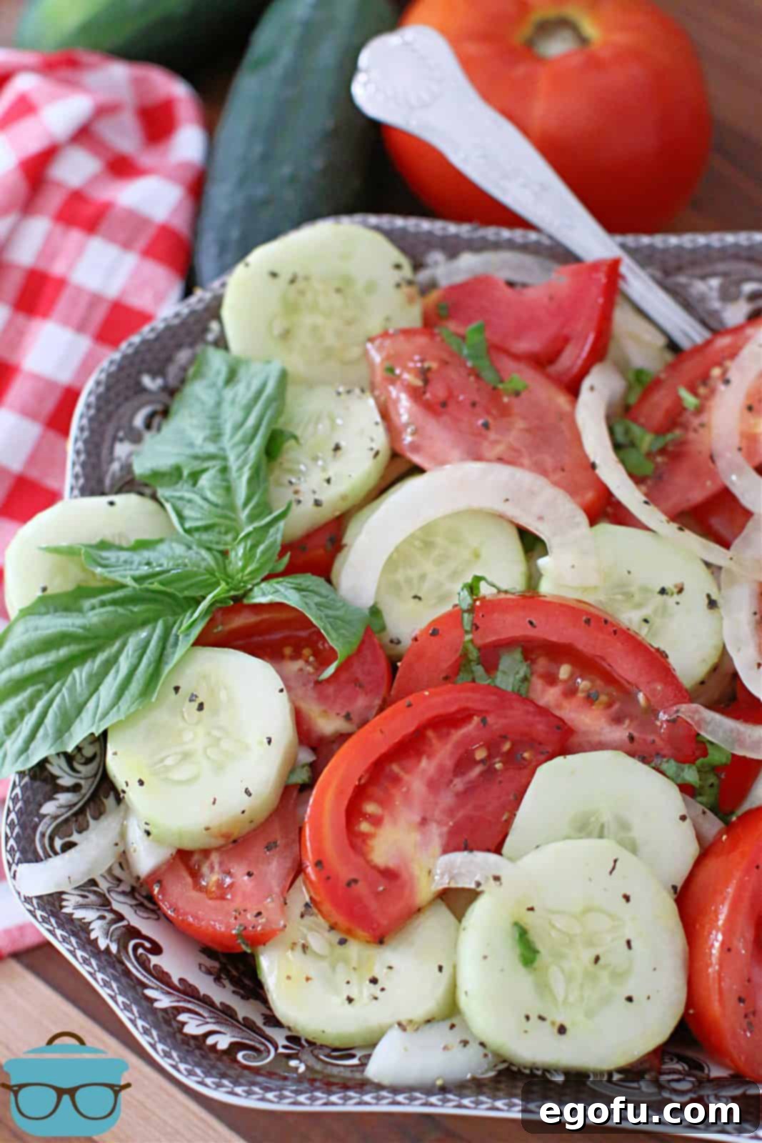 Marinated tomato, cucumber, and onion salad showcased in a beautiful brown and white Spode serving bowl with a spoon, accompanied by fresh whole cucumbers and a tomato in the background.
