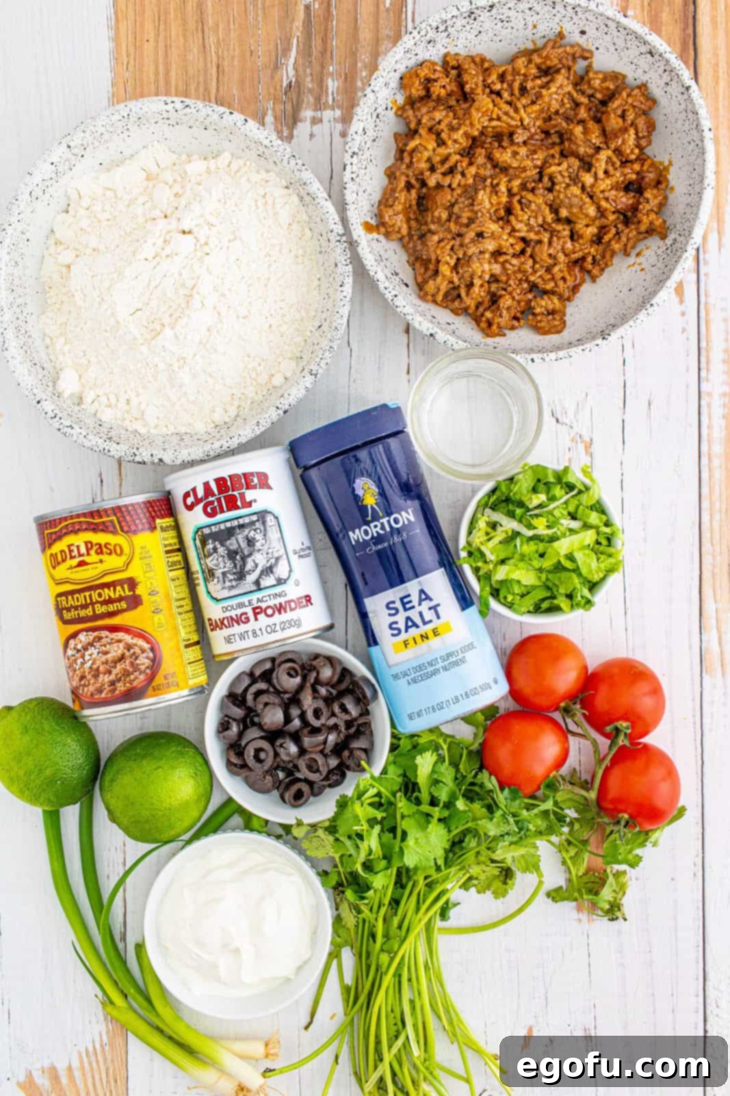 A collection of ingredients laid out: all-purpose flour, baking powder, fine sea salt, warm water, peanut oil, seasoned taco meat, refried beans, and various taco toppings.