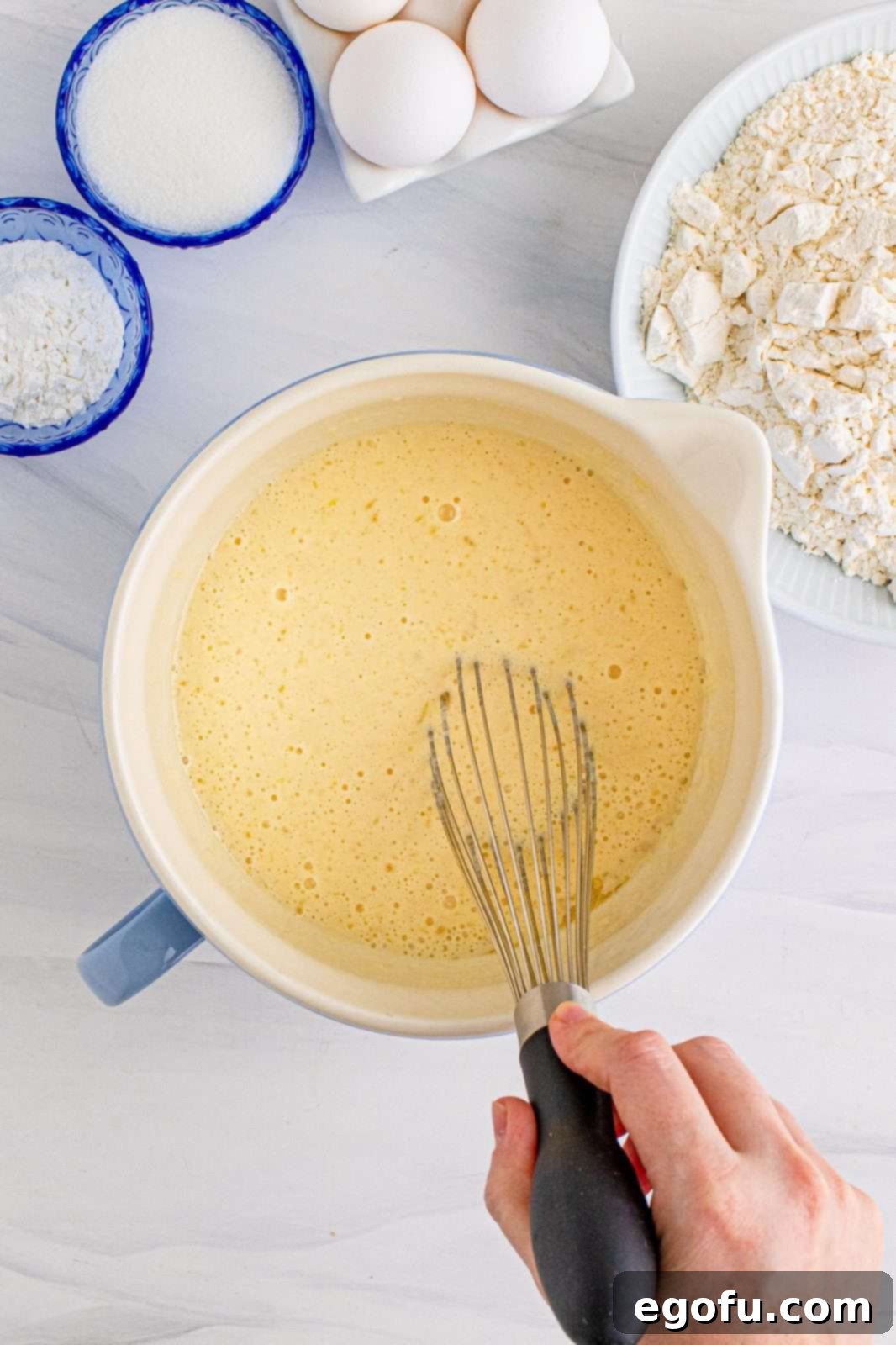 A whisk blending liquid ingredients—melted butter, buttermilk, eggs, and vanilla extract—in a white mixing bowl until smooth.