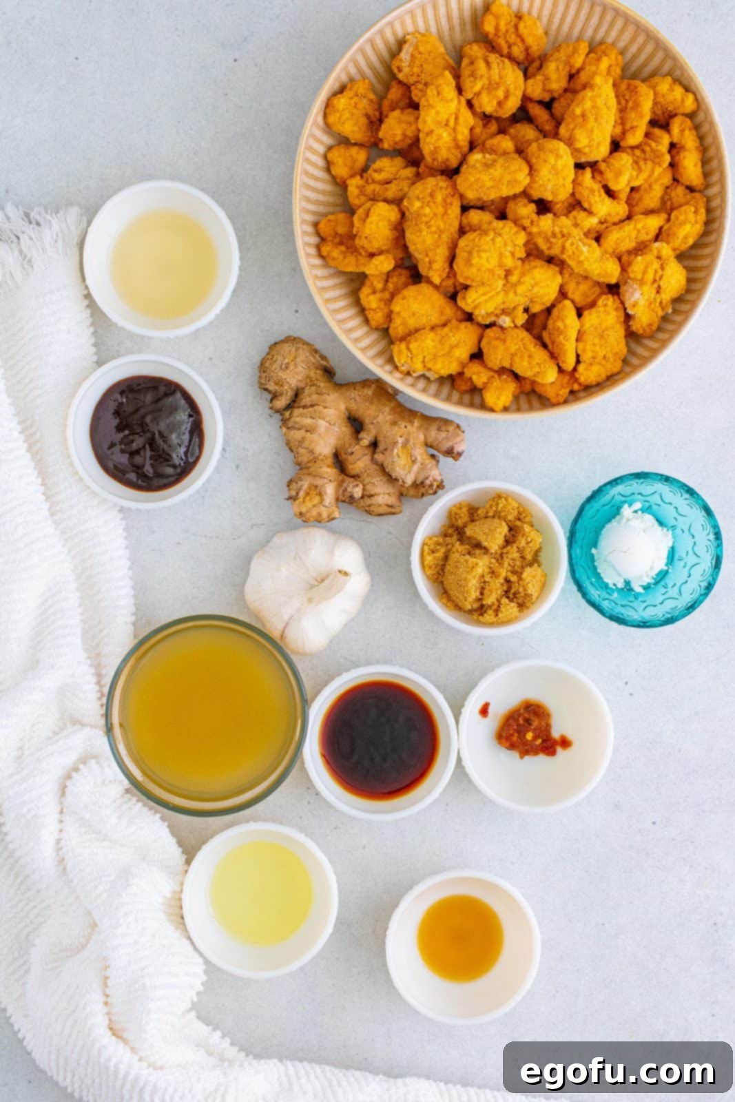 A vibrant collection of ingredients for General Tso's Chicken, including popcorn chicken, chicken stock, light brown sugar, soy sauce, hoisin sauce, rice wine vinegar, cornstarch, sesame oil, vegetable oil, minced garlic, fresh ginger, chili paste, scallions, and sesame seeds, neatly arranged.