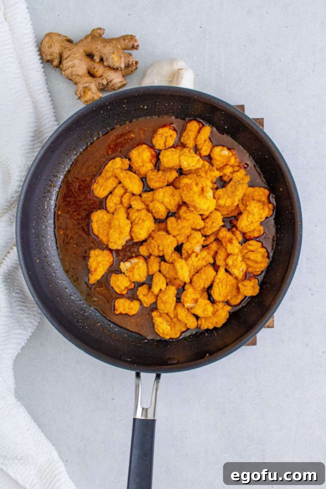 Cooked popcorn chicken pieces being added to the rich, thickened General Tso's sauce in a large skillet, ready to be tossed for even coating.