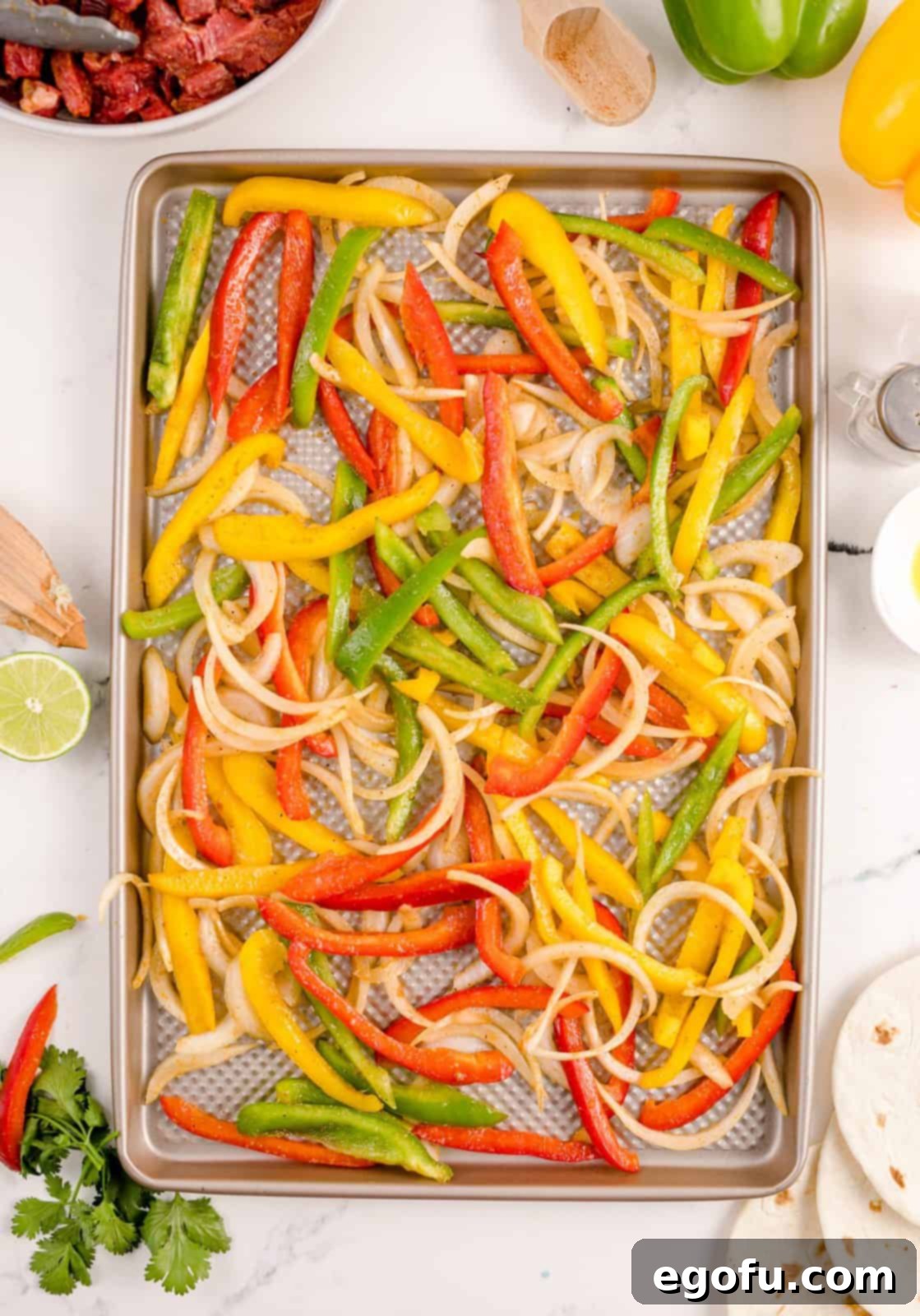 Thinly sliced bell peppers and onions spread in a single, even layer on a baking sheet, ready for the oven.