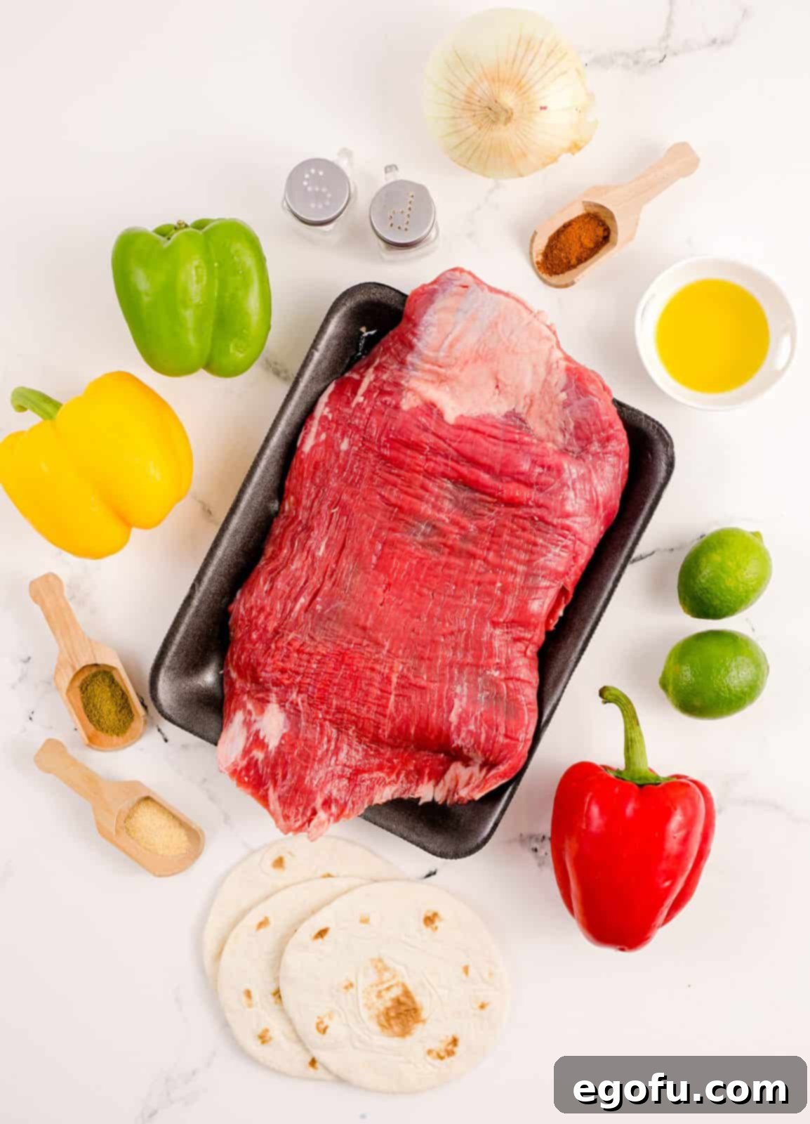 A flat lay image displaying all the fresh ingredients needed for Sheet Pan Beef Fajitas: flank steak, red, green, and yellow bell peppers, a yellow onion, olive oil, salt, pepper, cumin, chili powder, garlic powder, lime juice, and a stack of warm flour tortillas with optional garnishes like guacamole and sour cream.