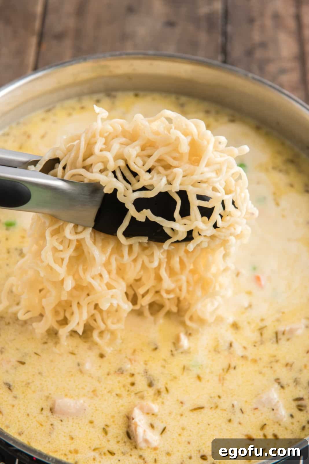 Ramen noodles being added to skillet with a pair of tongs, ready to be mixed into the creamy sauce.