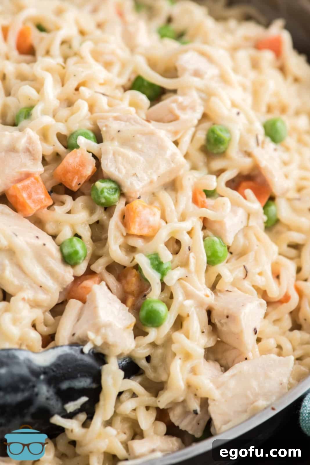 A close-up photo of a pair of tongs grasping some of the perfectly mixed and finished Chicken Pot Pie Ramen, highlighting its creamy texture.