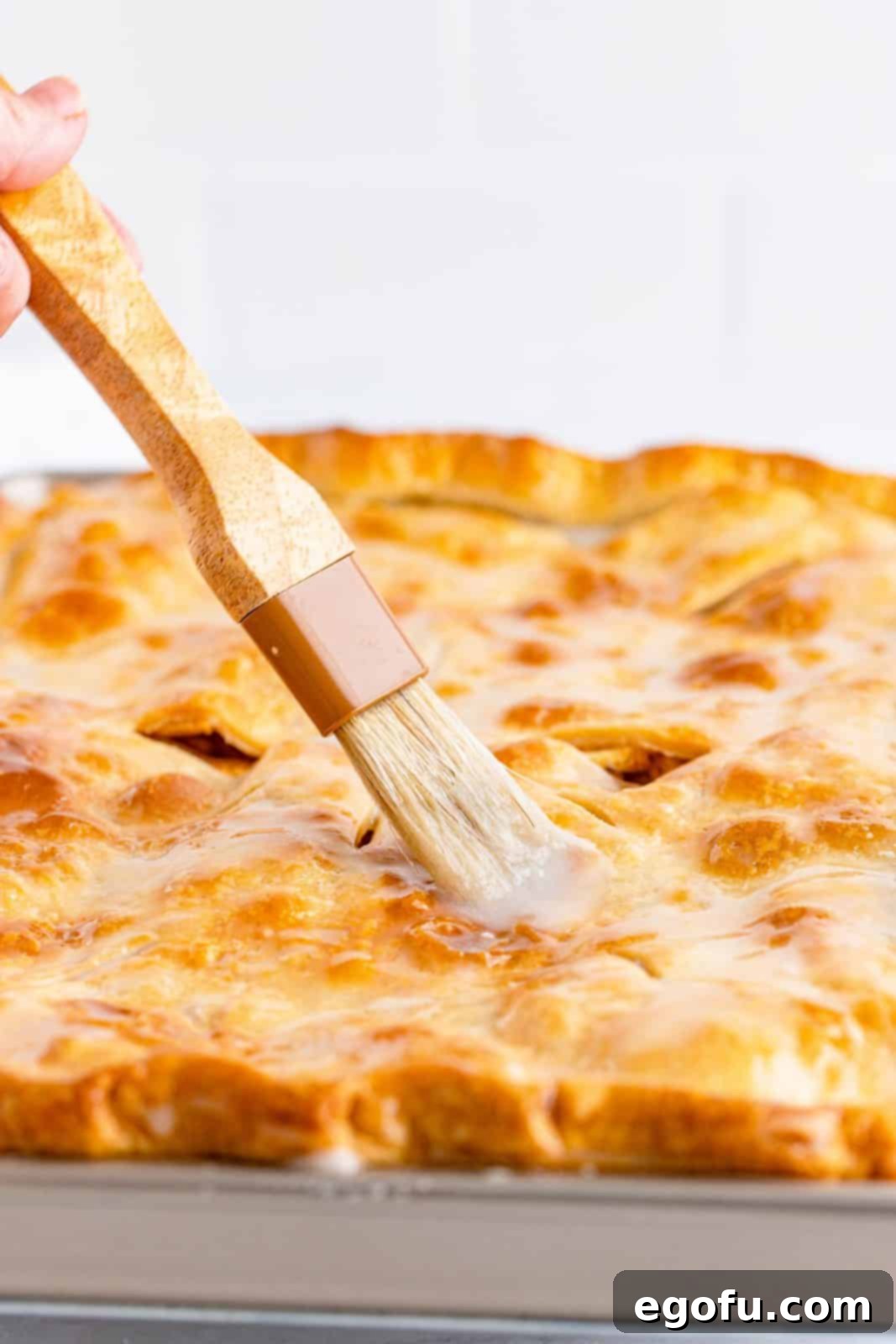 A pastry brush applying a smooth, white glaze to the top of the warm Apple Slab Pie.