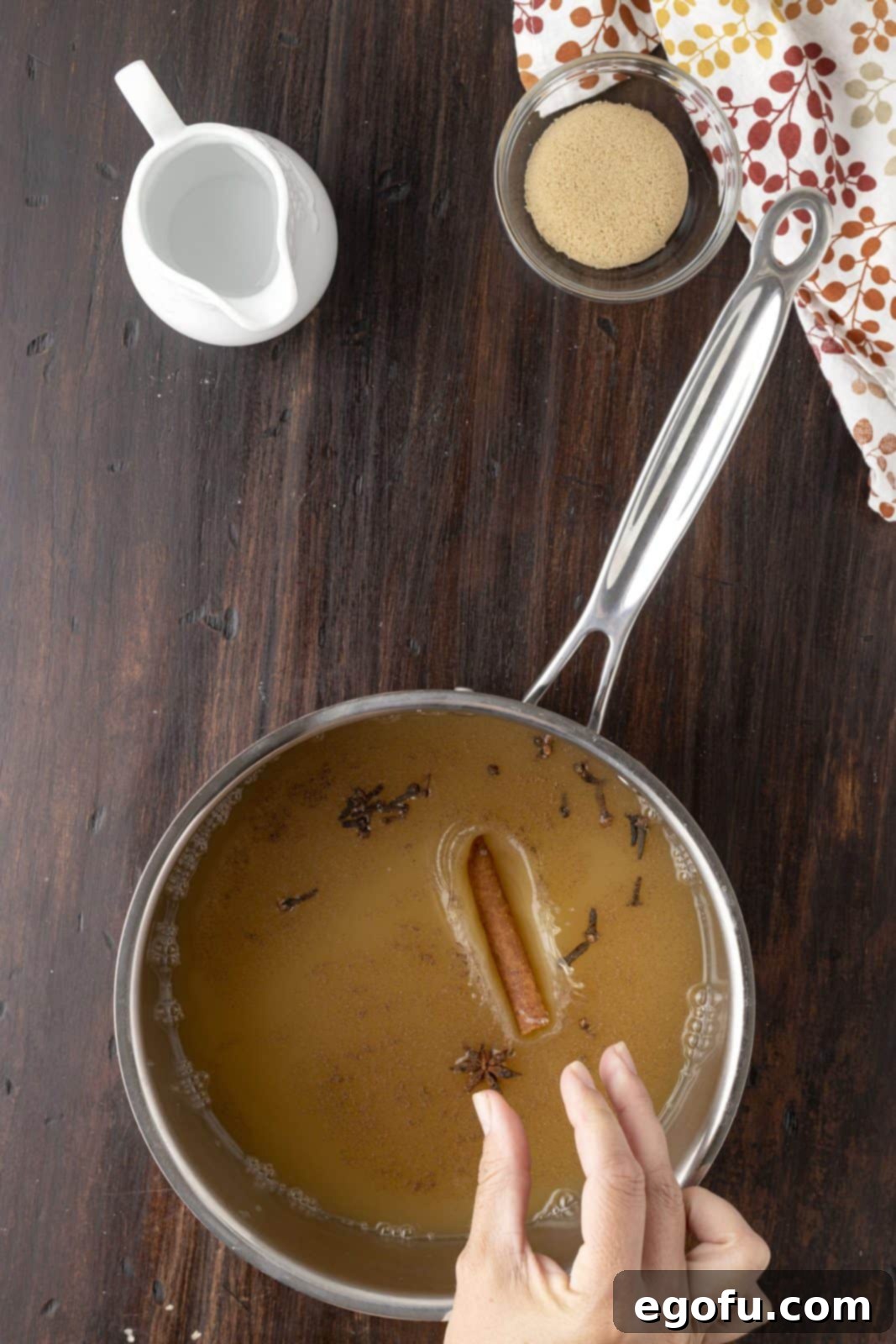 Apple cider, brown sugar, cinnamon stick, star anise, whole cloves, and allspice being added to a saucepan on a stovetop.