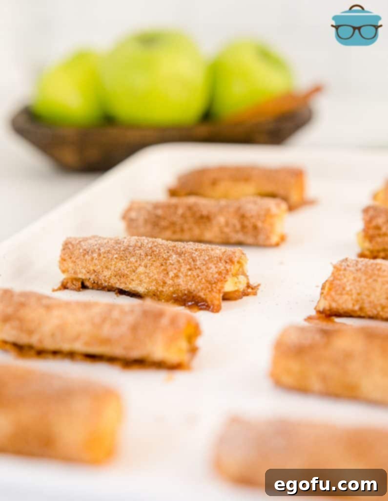 A perfectly arranged display of finished Baked Apple Pie Rollups on a white board, with fresh apples blurred in the background, highlighting the inviting warmth of the baked treats.