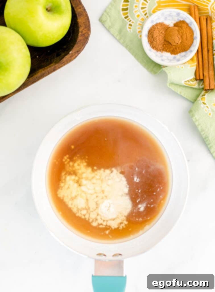 A white saucepan on a stovetop, filled with the liquid ingredients for the apple pie filling: water, brown sugar, maple syrup, caramel syrup, lemon juice, and cornstarch, all whisked together.