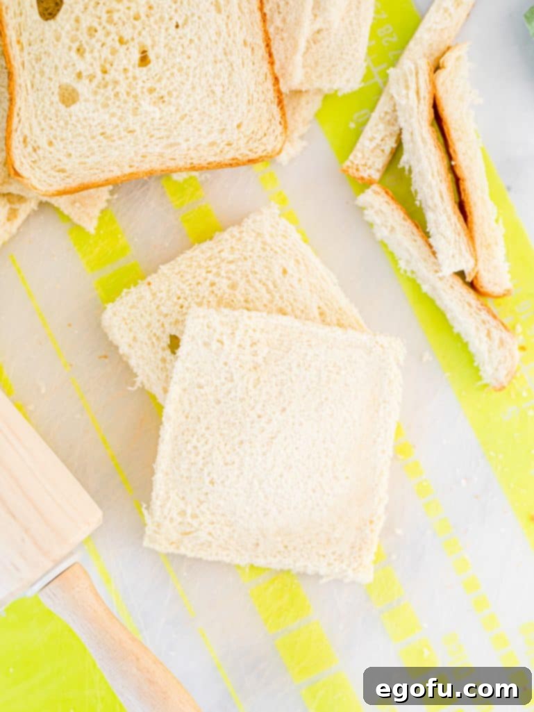Several slices of sandwich bread with their crusts neatly removed, flattened with a rolling pin on a cutting board, ready for filling.