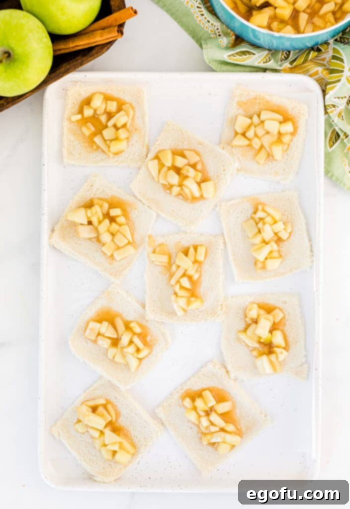 A flattened slice of sandwich bread with a generous dollop of apple pie filling placed precisely in the center, awaiting rolling.