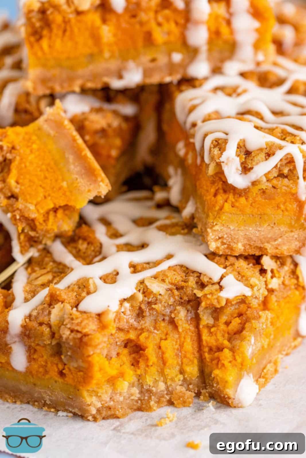 Close-up photo of pumpkin streusel bars stacked on white parchment paper, with a bite removed from one bar, showing the layers and drizzle.