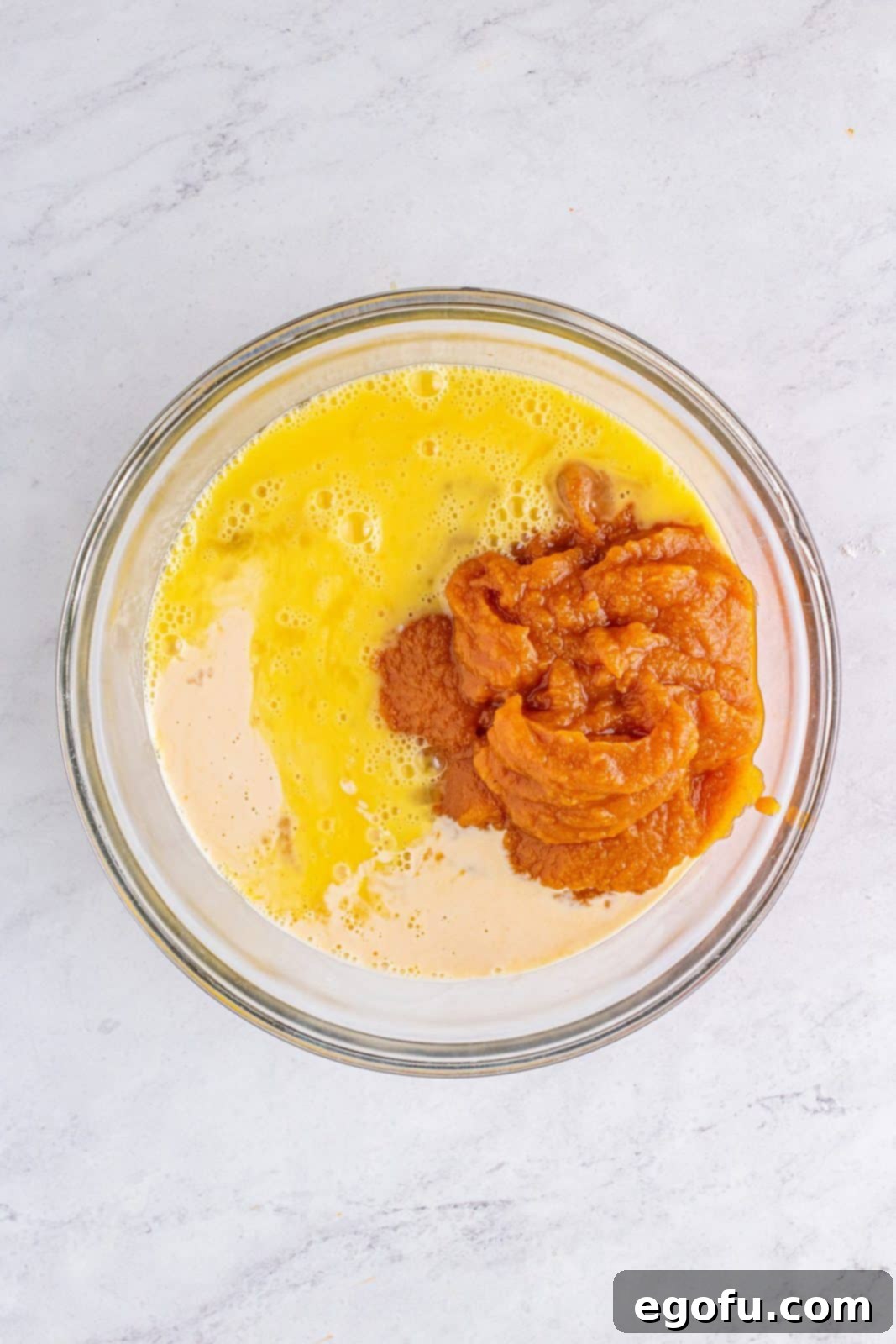 Pumpkin mix, beaten eggs, and evaporated milk are added to a clear bowl, ready to be whisked for the filling.