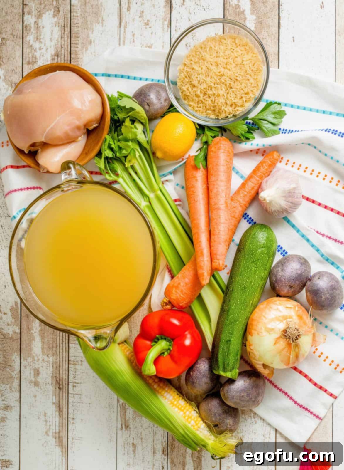 Golden Harvest Chicken and Rice Soup 4 A vibrant display of fresh ingredients for Harvest Chicken and Rice Soup, including salted butter, a whole onion, carrots, celery, red pepper, garlic cloves, chicken stock cartons, purple potatoes, zucchini, an ear of corn, raw chicken breasts, uncooked white rice, bay leaves, and a fresh lemon.