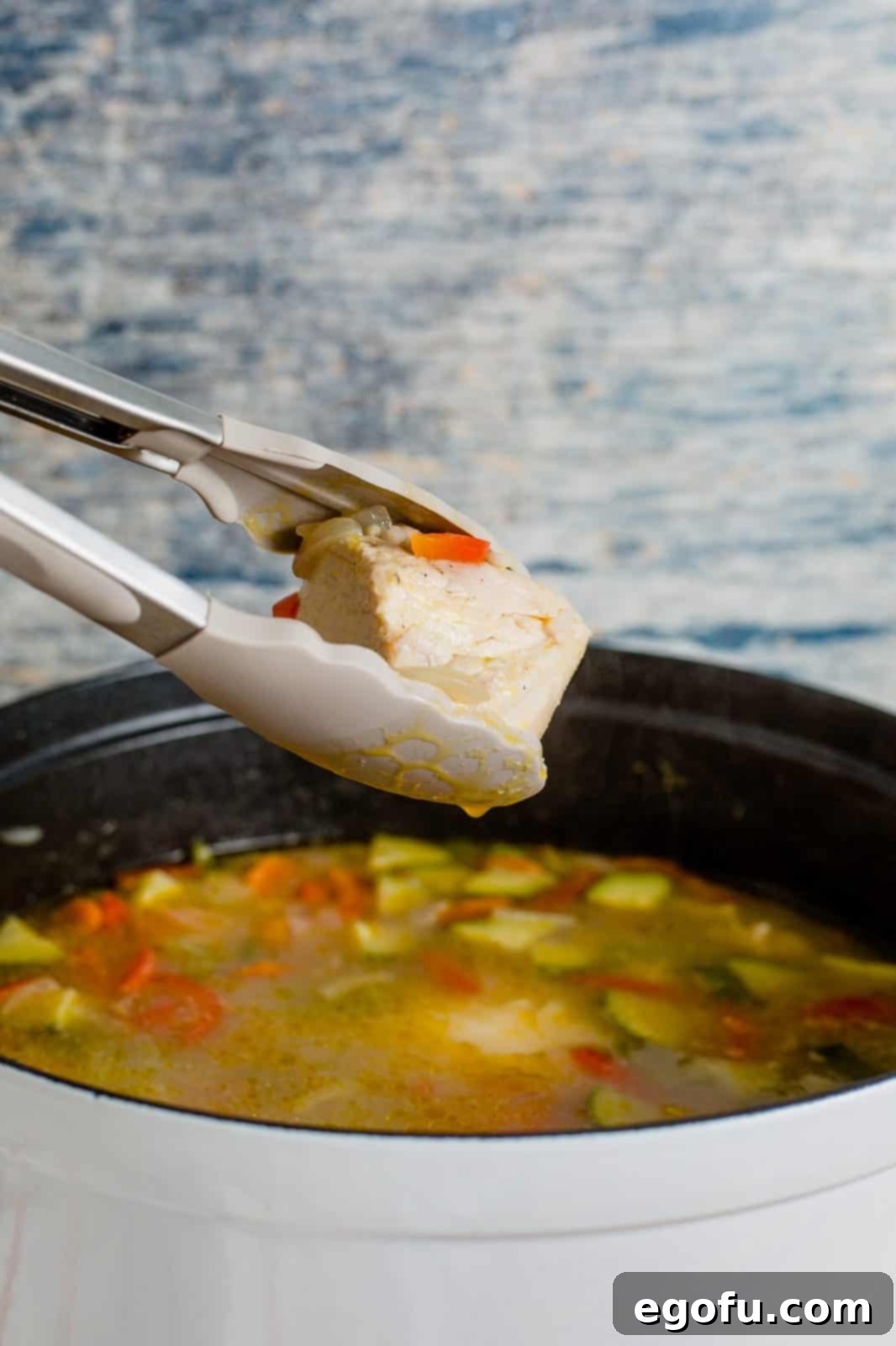 Golden Harvest Chicken and Rice Soup 8 Tongs lifting a cooked chicken breast out of the simmering soup in a pot, steam rising.