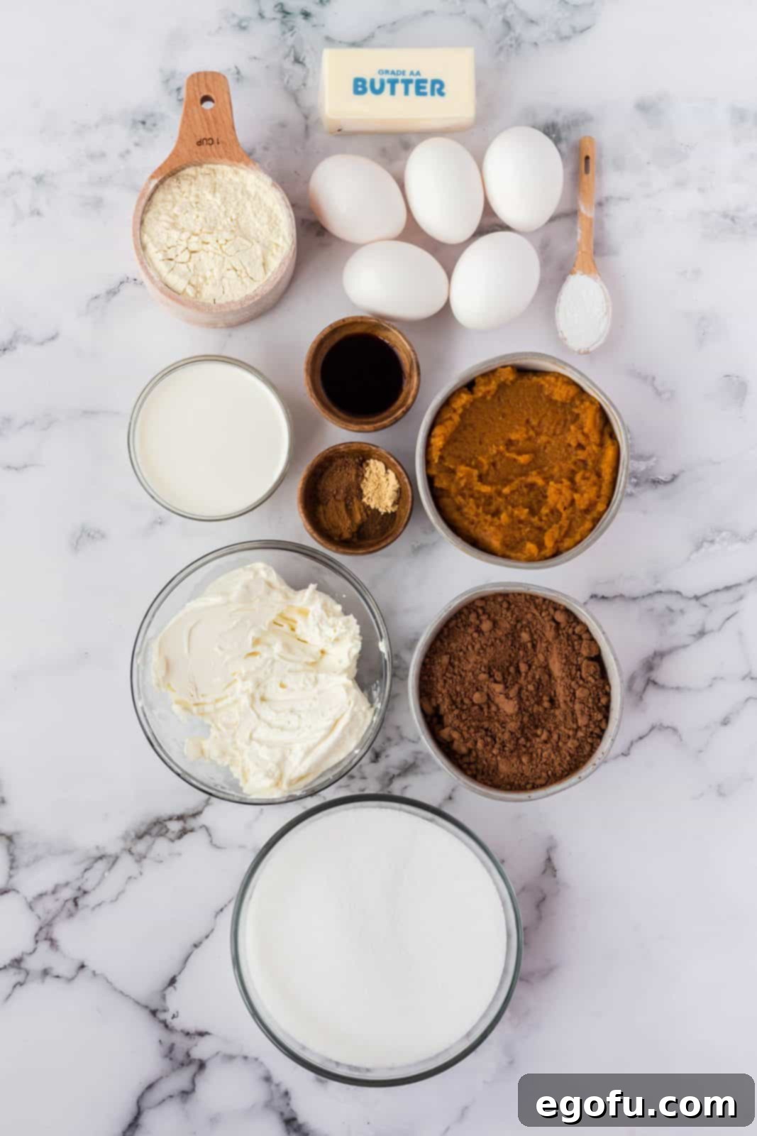 A flat lay photograph displaying all the ingredients needed for Pumpkin Swirl Brownies: Salted butter, granulated sugar, cocoa powder, eggs, vanilla extract, all-purpose flour, baking powder, cream cheese, pumpkin puree, ground cinnamon, ground ginger, ground nutmeg, ground cloves, and heavy whipping cream.
