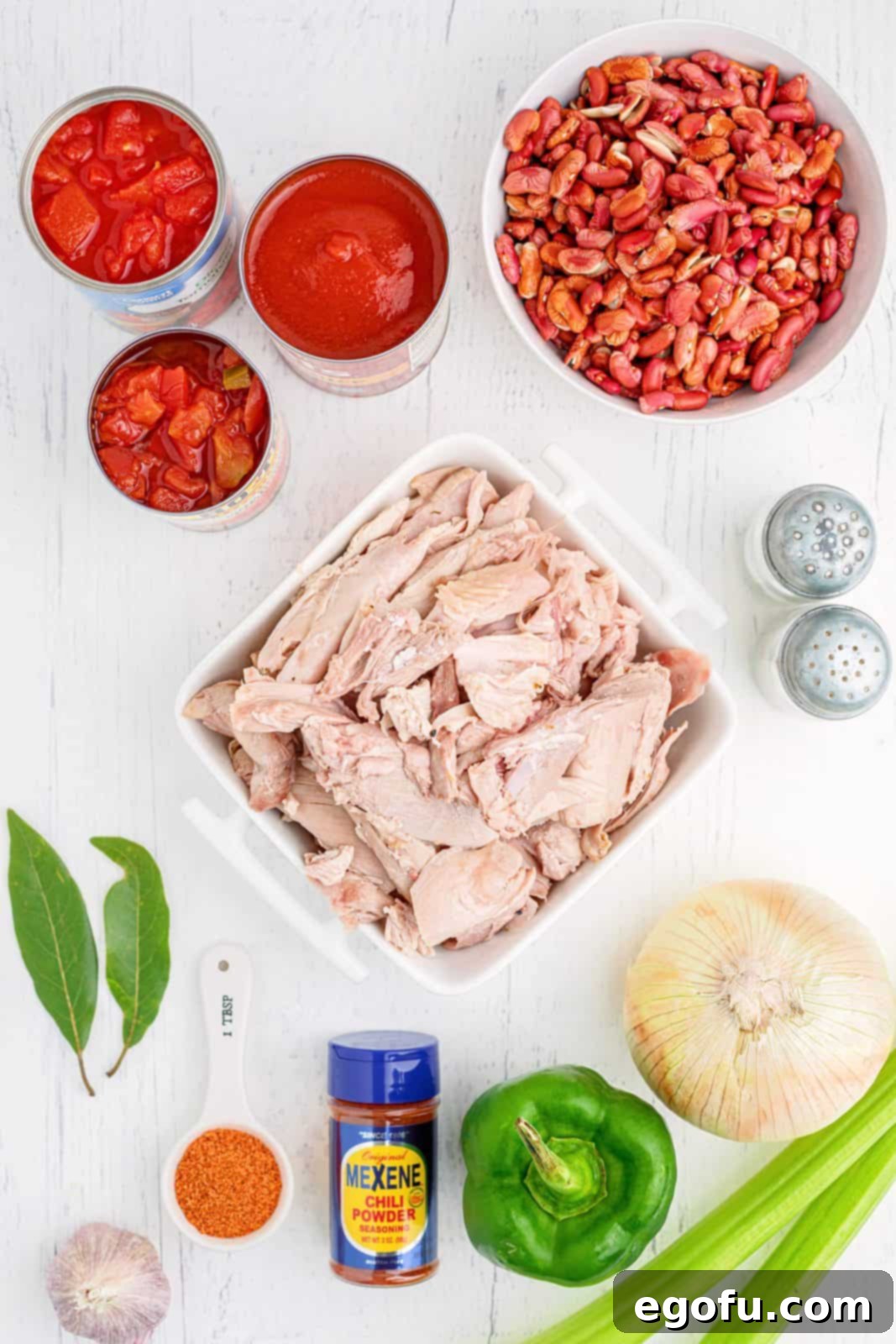 A colorful array of fresh and canned ingredients needed for Crock Pot Turkey Chili: leftover roast turkey, dry red kidney beans, chopped tomatoes and chiles, tomato sauce, chopped tomatoes, yellow onions, green bell pepper, celery ribs, garlic, bay leaves, creole seasoning, Mexene Chili Seasoning mix, salt, and pepper.