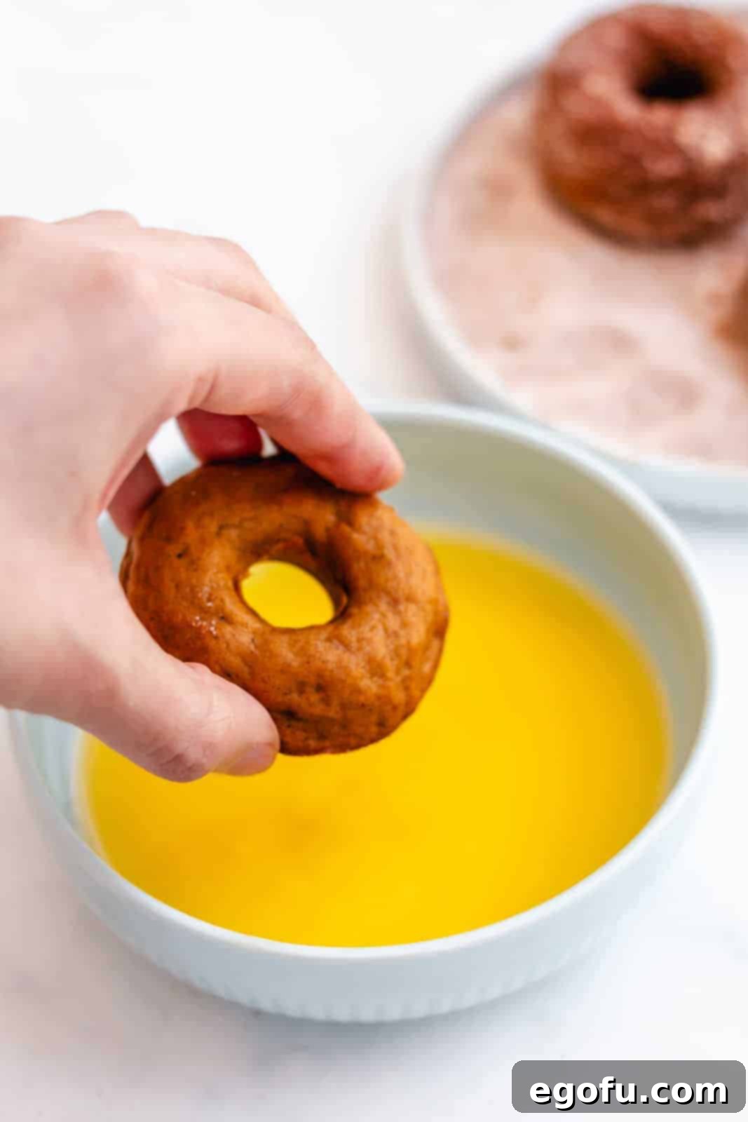 A hand dipping a baked pumpkin donut into a bowl of melted butter, preparing it for the cinnamon sugar coating.