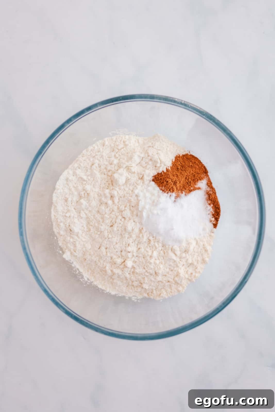Dry ingredients like flour, baking powder, baking soda, salt, cinnamon, and pumpkin pie spice added to a clear mixing bowl, ready to be whisked.