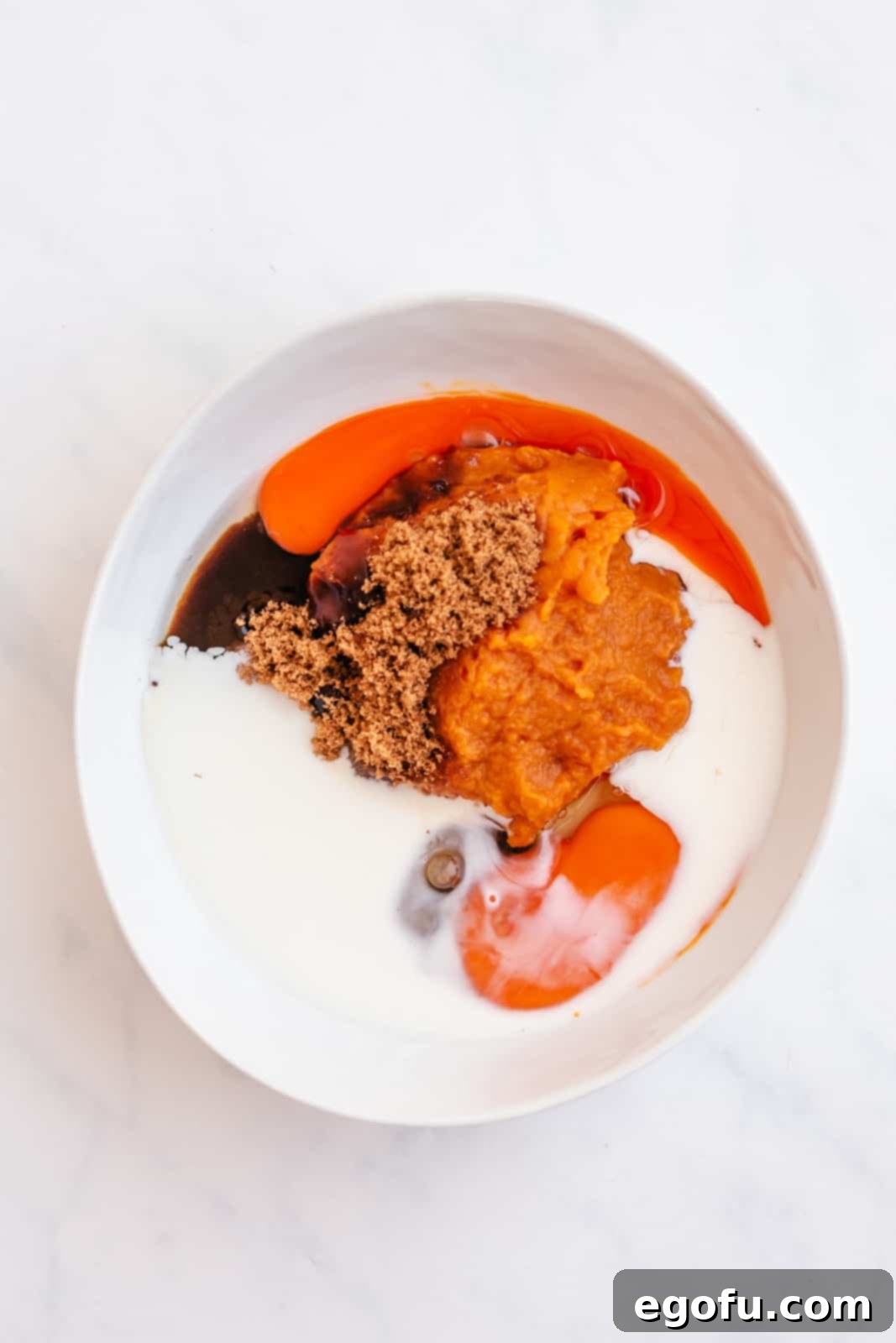 Wet ingredients including oil, brown sugar, eggs, pumpkin puree, milk, and vanilla extract, along with spices, combined in a white bowl and whisked.