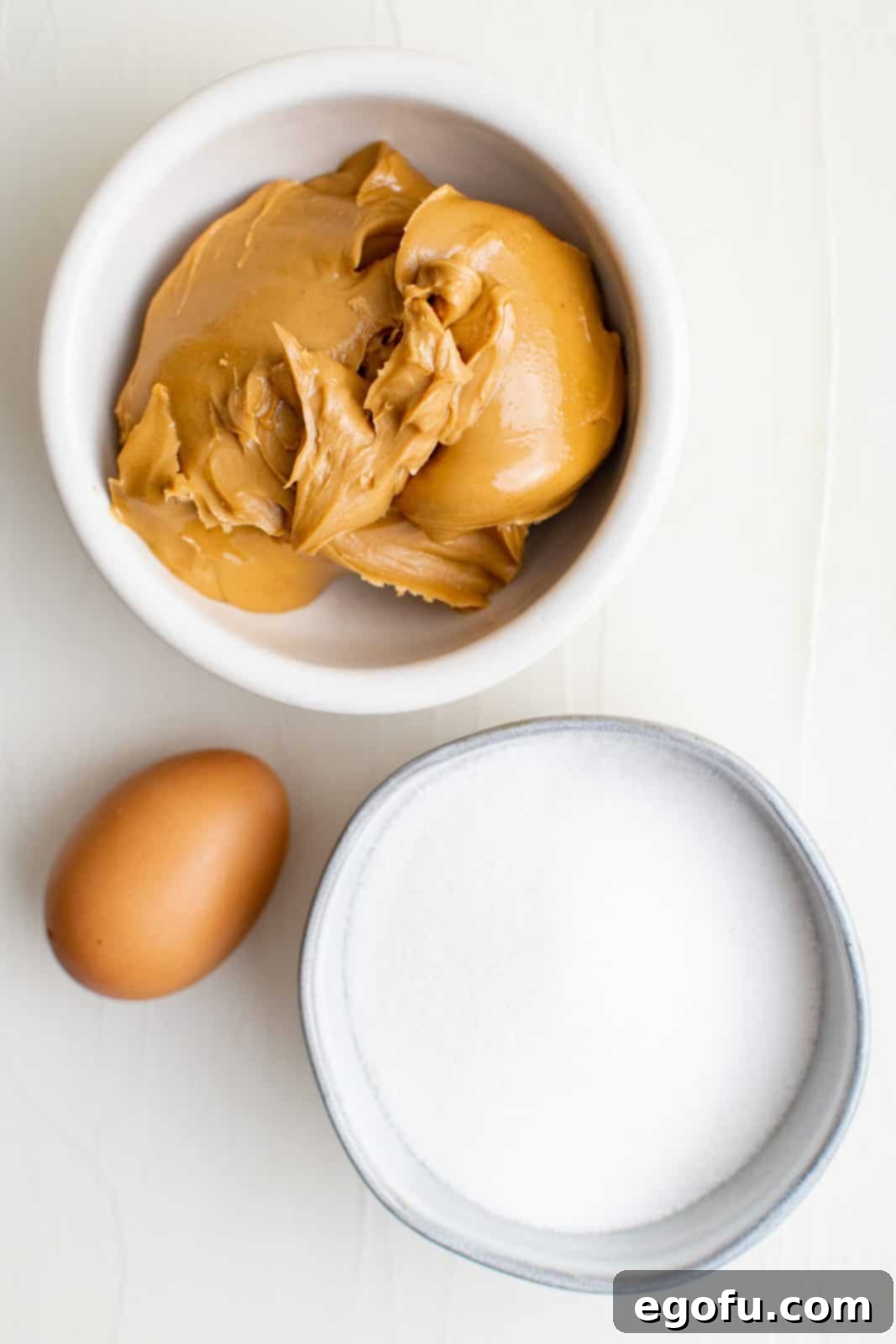 A clear photo showing the three essential ingredients: a jar of peanut butter, a bowl of granulated sugar, and a single egg, ready for baking.