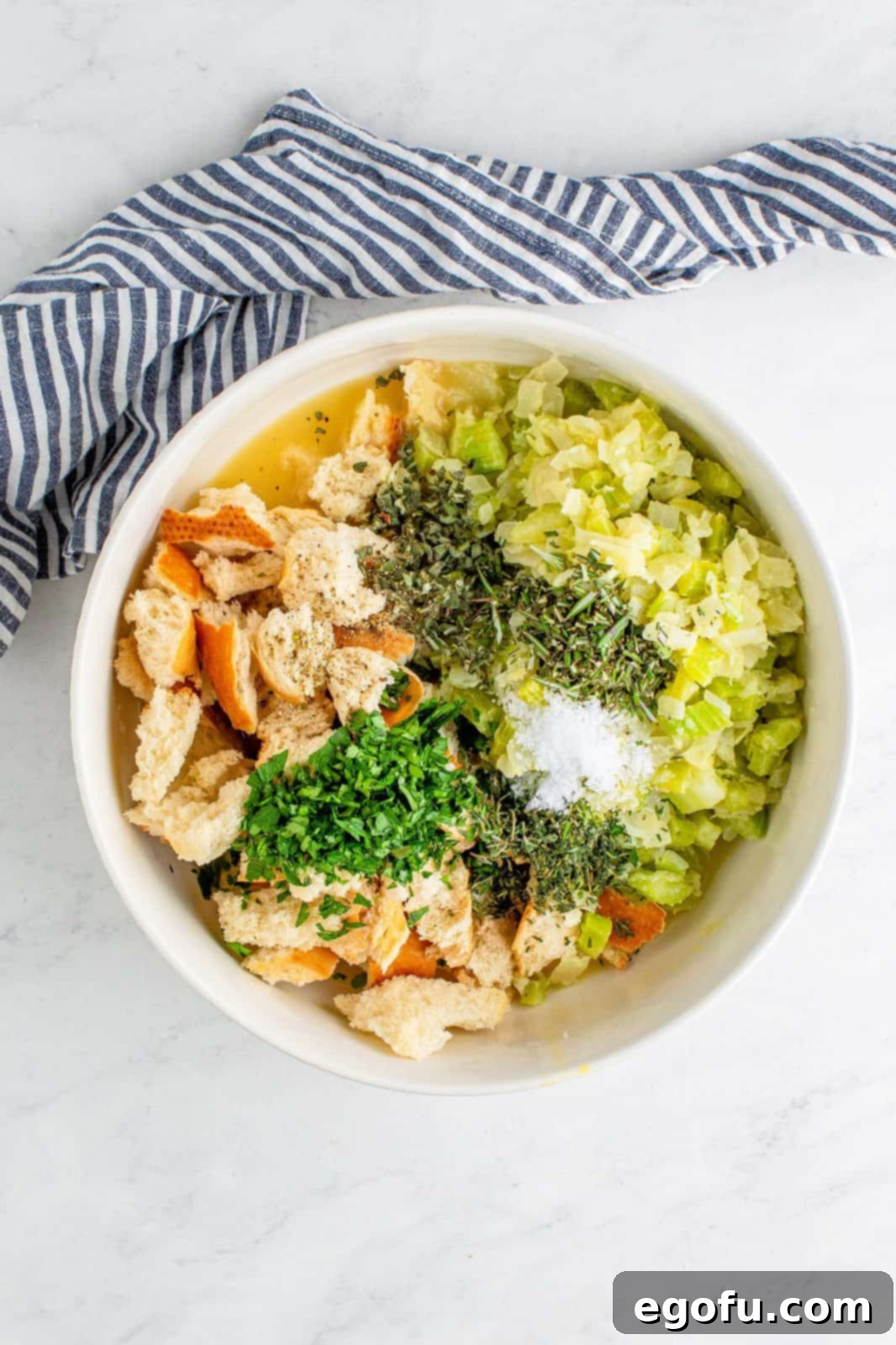 All stuffing ingredients, including dried bread, fresh herbs, sautéed vegetables, and the egg-stock mixture, tossed together in a large mixing bowl.