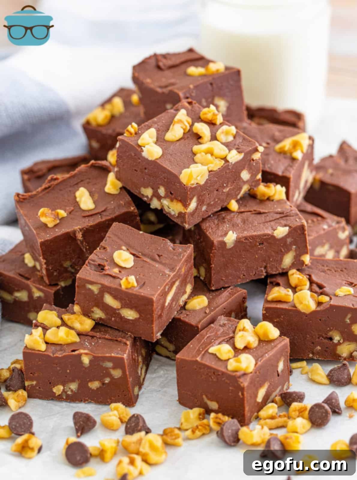 Stacked squares of Chocolate Walnut Fudge with whole walnuts and chocolate chips scattered at the bottom, highlighting the finished product.