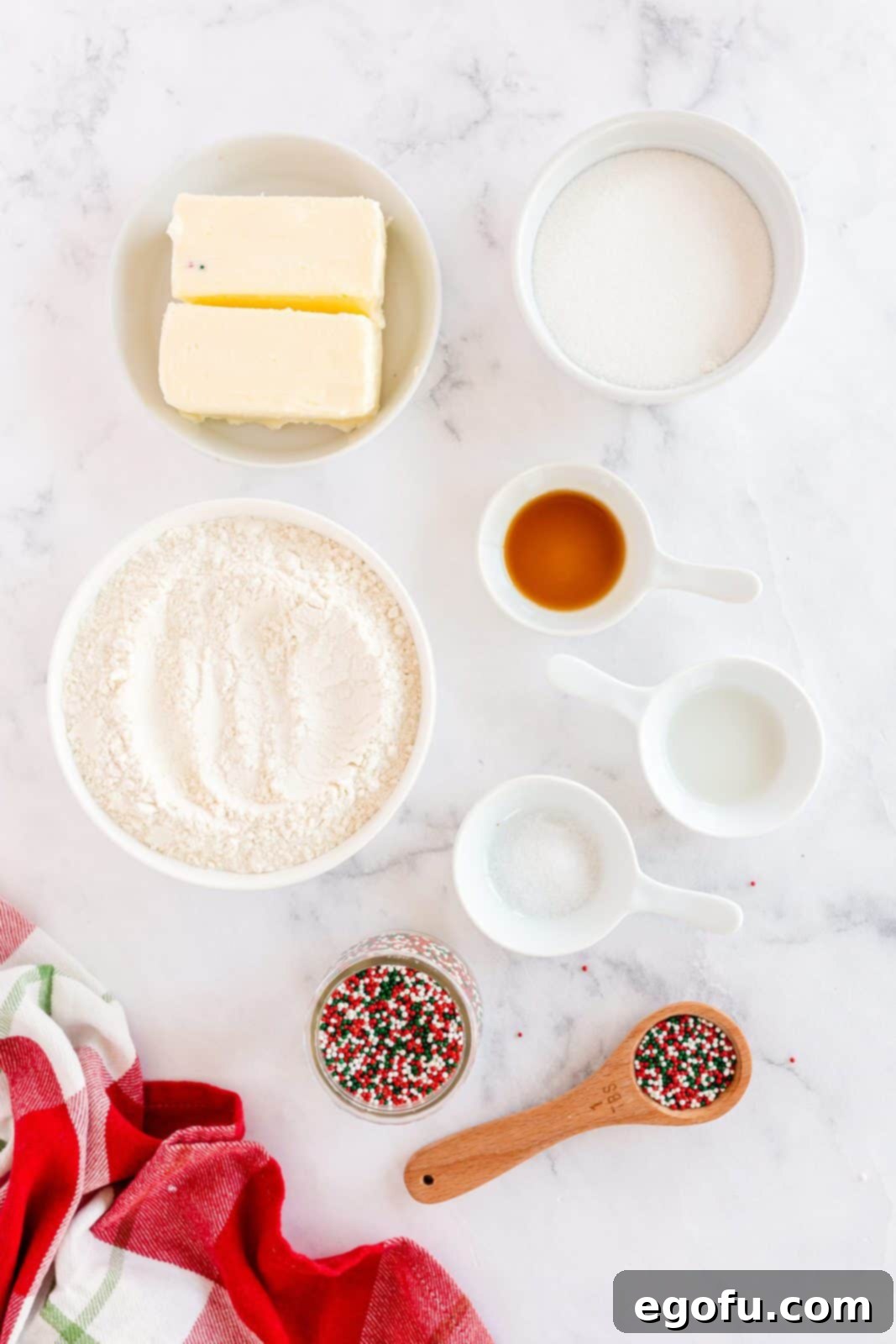 A flat lay of the ingredients needed for Mini Christmas Shortbread Cookies: salted butter, granulated sugar, vanilla extract, almond extract, all-purpose flour, and nonpareils.
