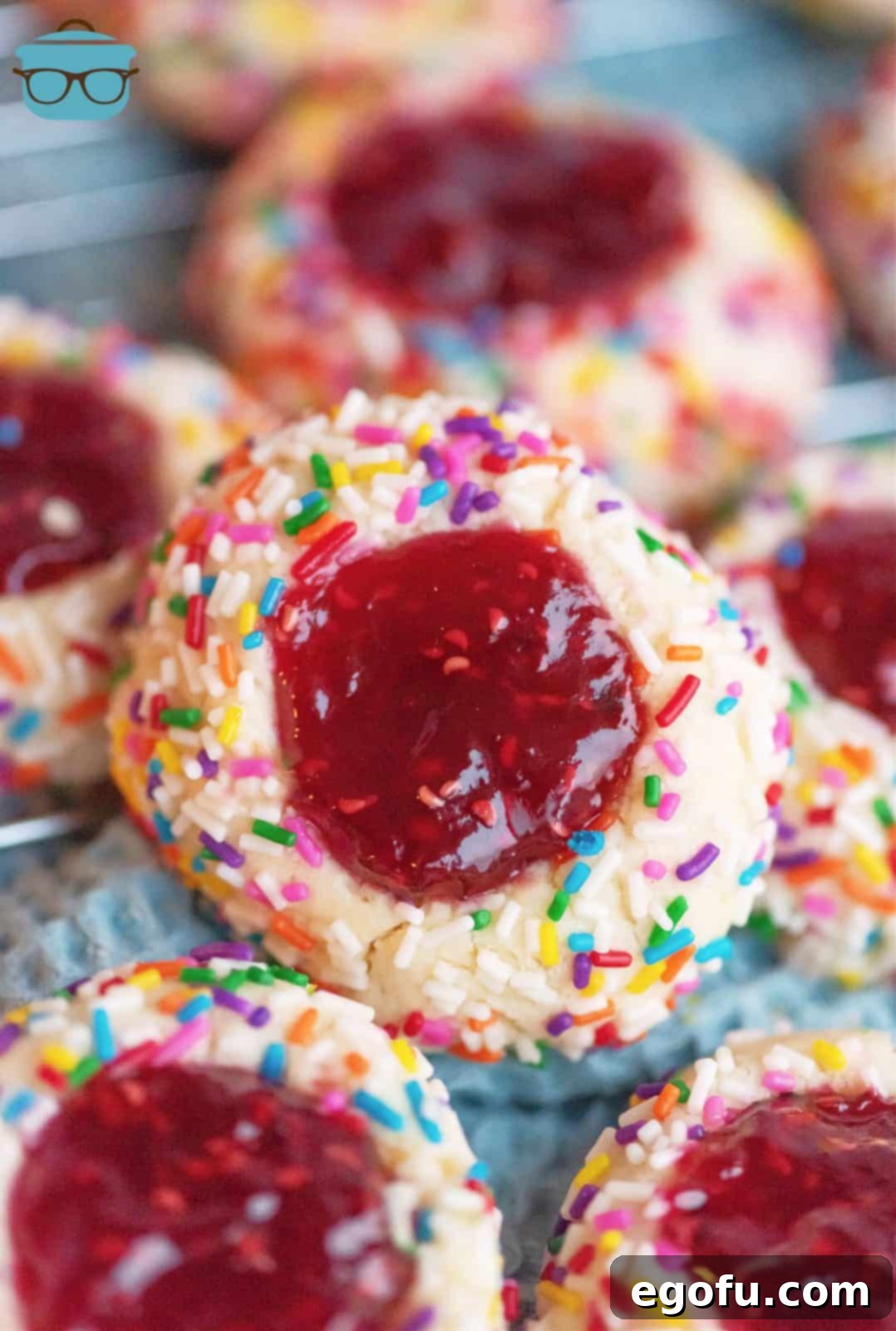 Raspberry Thumbprint Cookies on a rustic board, with one cookie stacked showing its vibrant jam filling and sprinkle-coated edge.