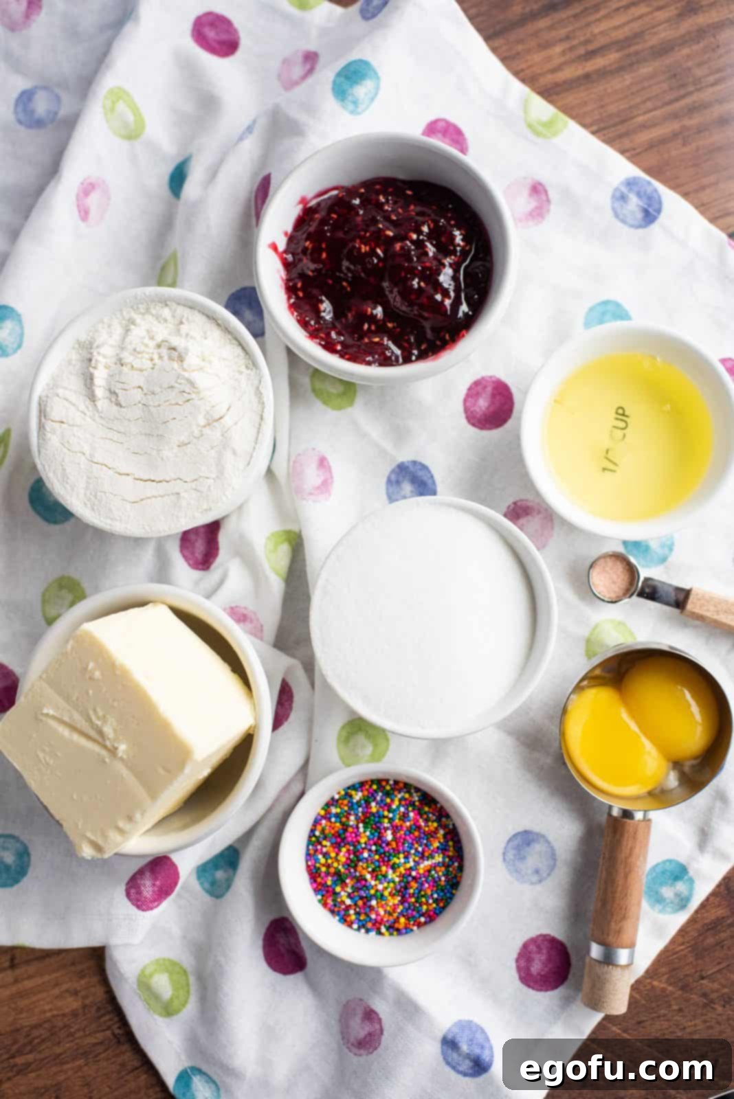A flat lay photograph showing all the essential ingredients for Raspberry Thumbprint Cookies: butter, granulated sugar, egg yolks, salt, all-purpose flour, sprinkles, and raspberry jam.