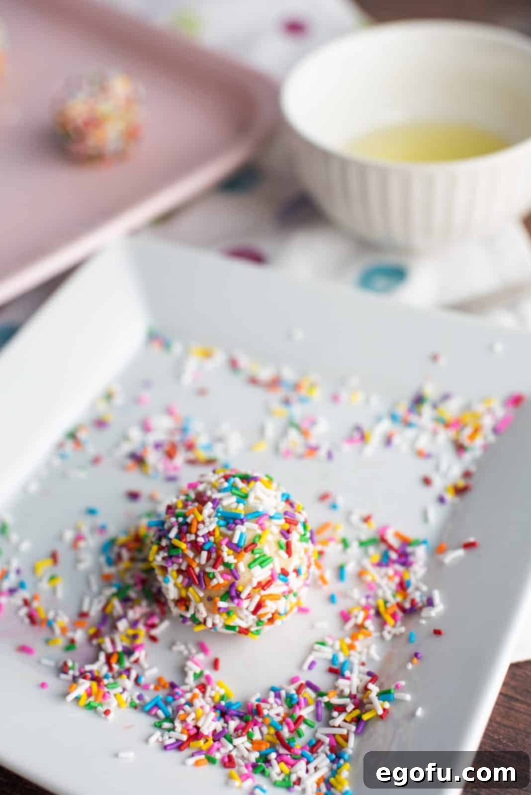Cookie dough balls being rolled in a bowl filled with colorful sprinkles for a festive coating.
