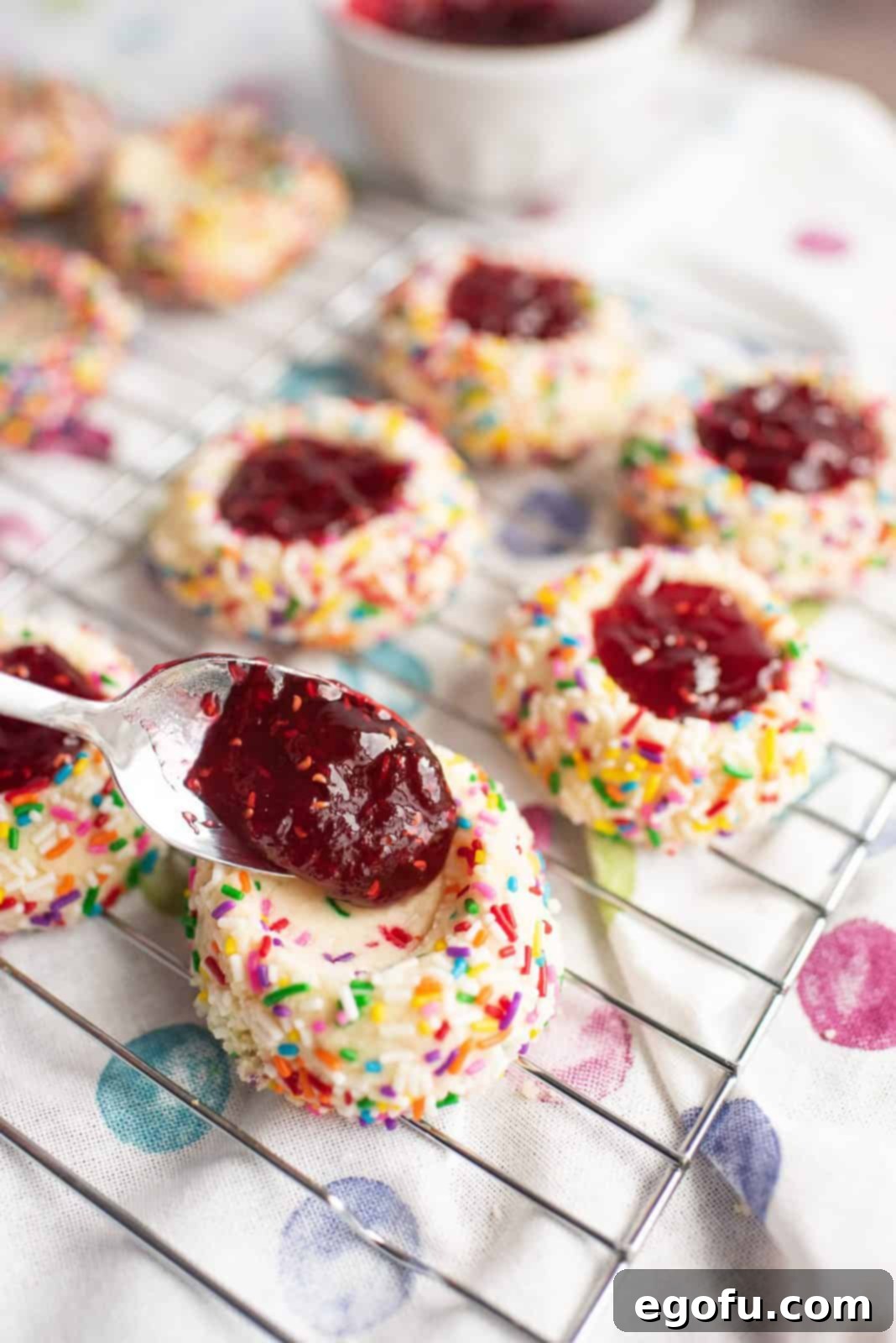 Raspberry jam being carefully filled into the indentations of each thumbprint cookie on a baking sheet.
