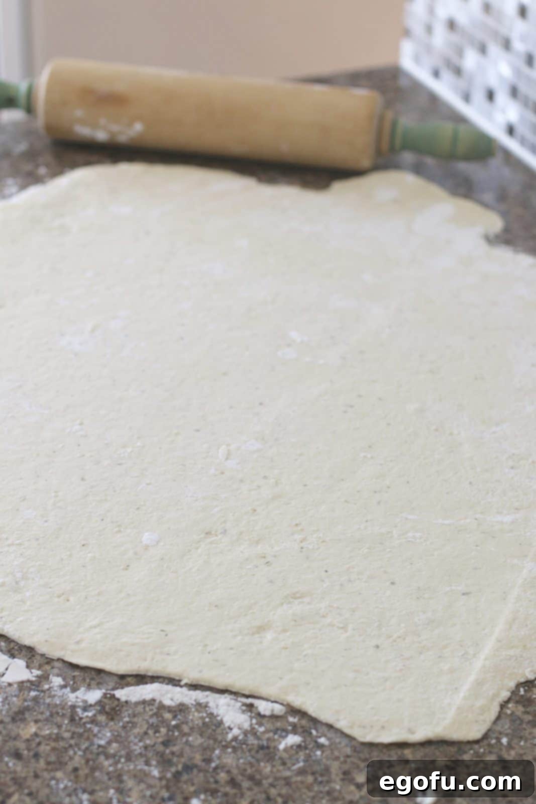 Flat, rolled out pizza dough on a countertop with a wooden rolling pin beside it.
