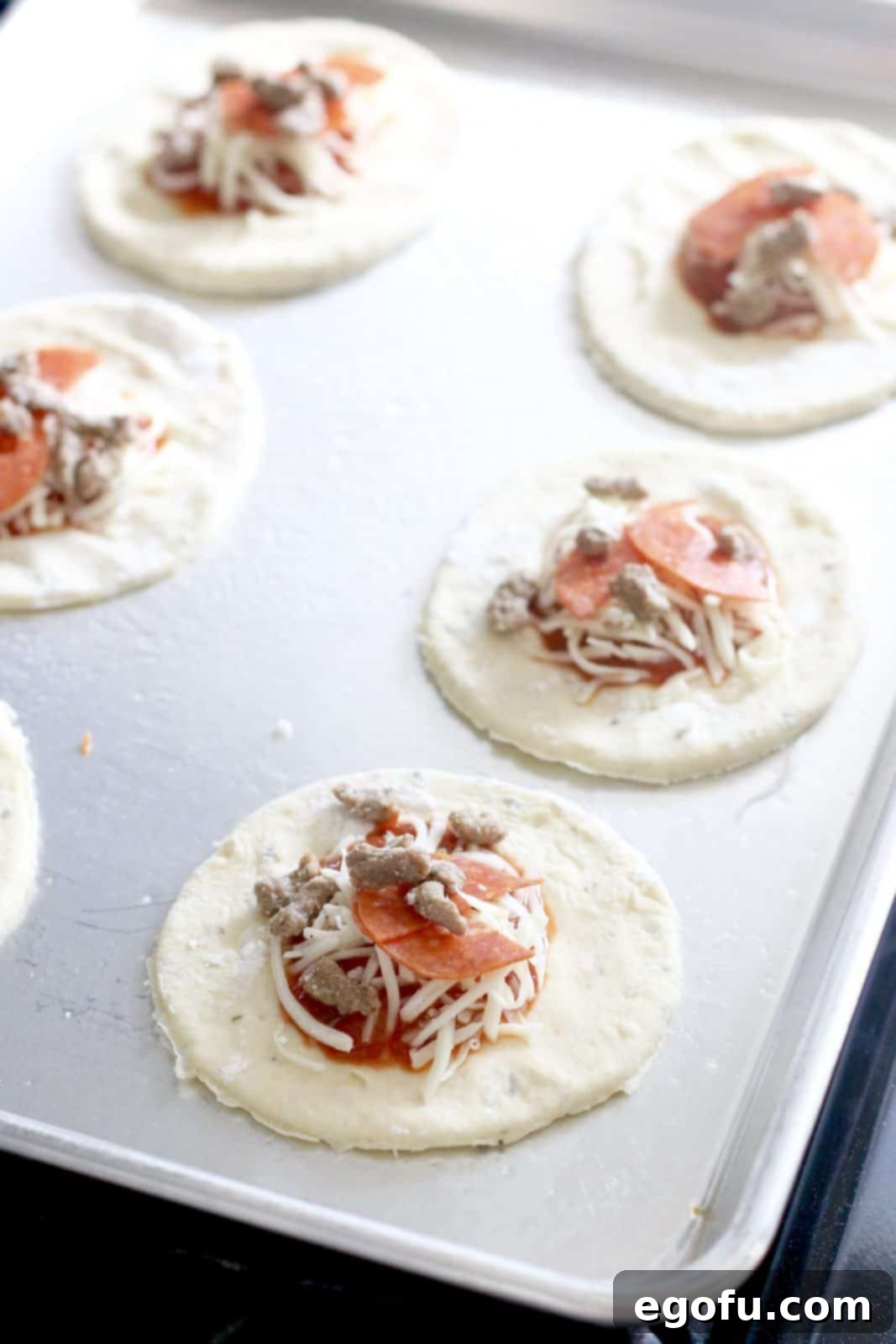 Pizza sauce, shredded cheese, and crumbled sausage are carefully placed into the middle of each pizza dough circle on a baking sheet.
