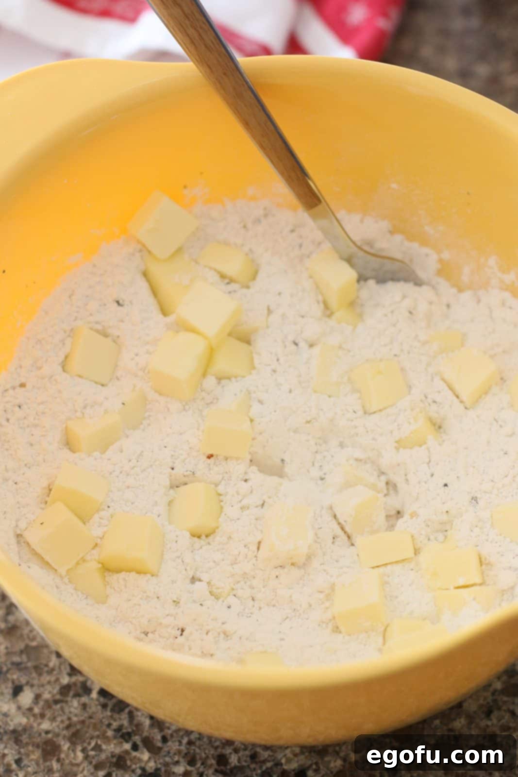 Cubed cold salted butter is being cut into the flour mixture with a fork in a yellow mixing bowl.