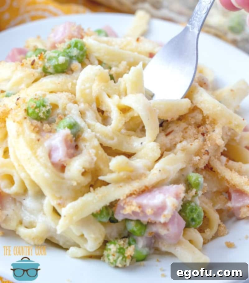 A fork lifts a generous portion of creamy ham fettuccine casserole from a white plate, showcasing the rich texture and delicious ingredients.