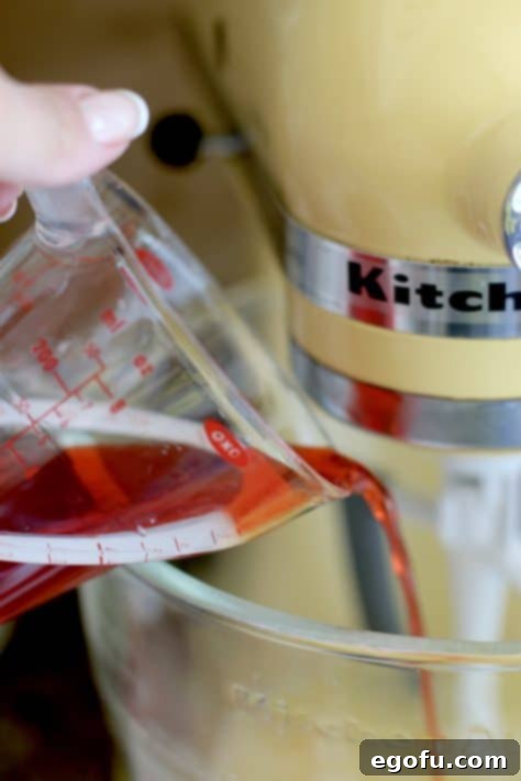 A close-up shot showing maraschino cherry juice being poured into a yellow KitchenAid stand mixer bowl filled with other cake mix ingredients.