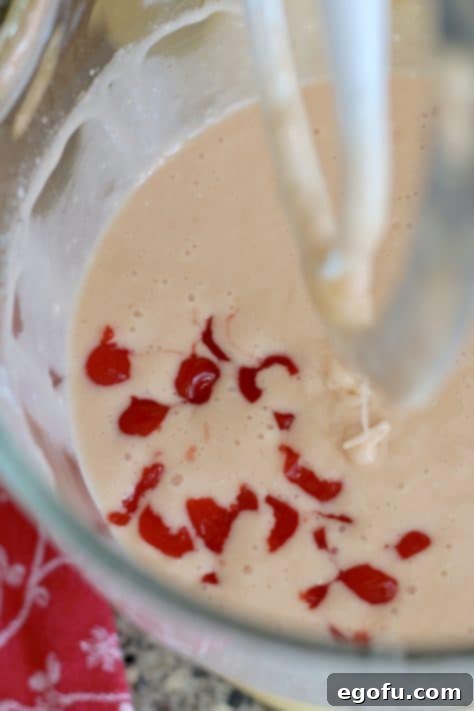 Chopped maraschino cherries being gently stirred into the pink cake batter in a mixing bowl with a spatula.