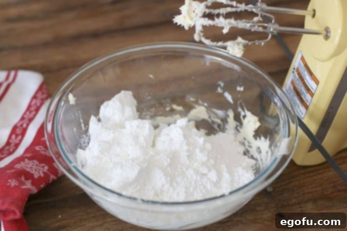 Powdered sugar being slowly added to the creamed butter and cream cheese mixture in a bowl, preventing a mess.