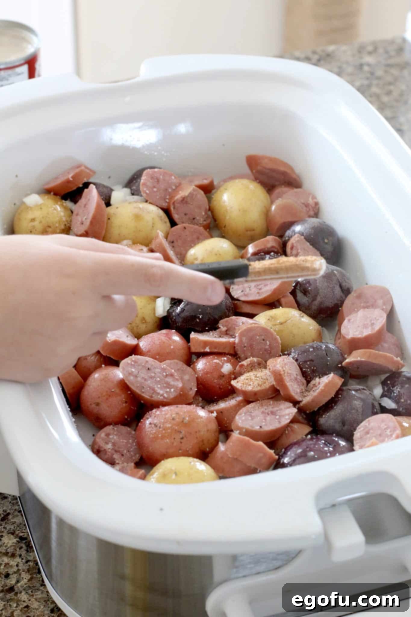 Easy Slow Cooker Creamy Sausage and Potatoes 6 Cajun seasoning being sprinkled over a mix of potatoes, sausage, and onions in a slow cooker.