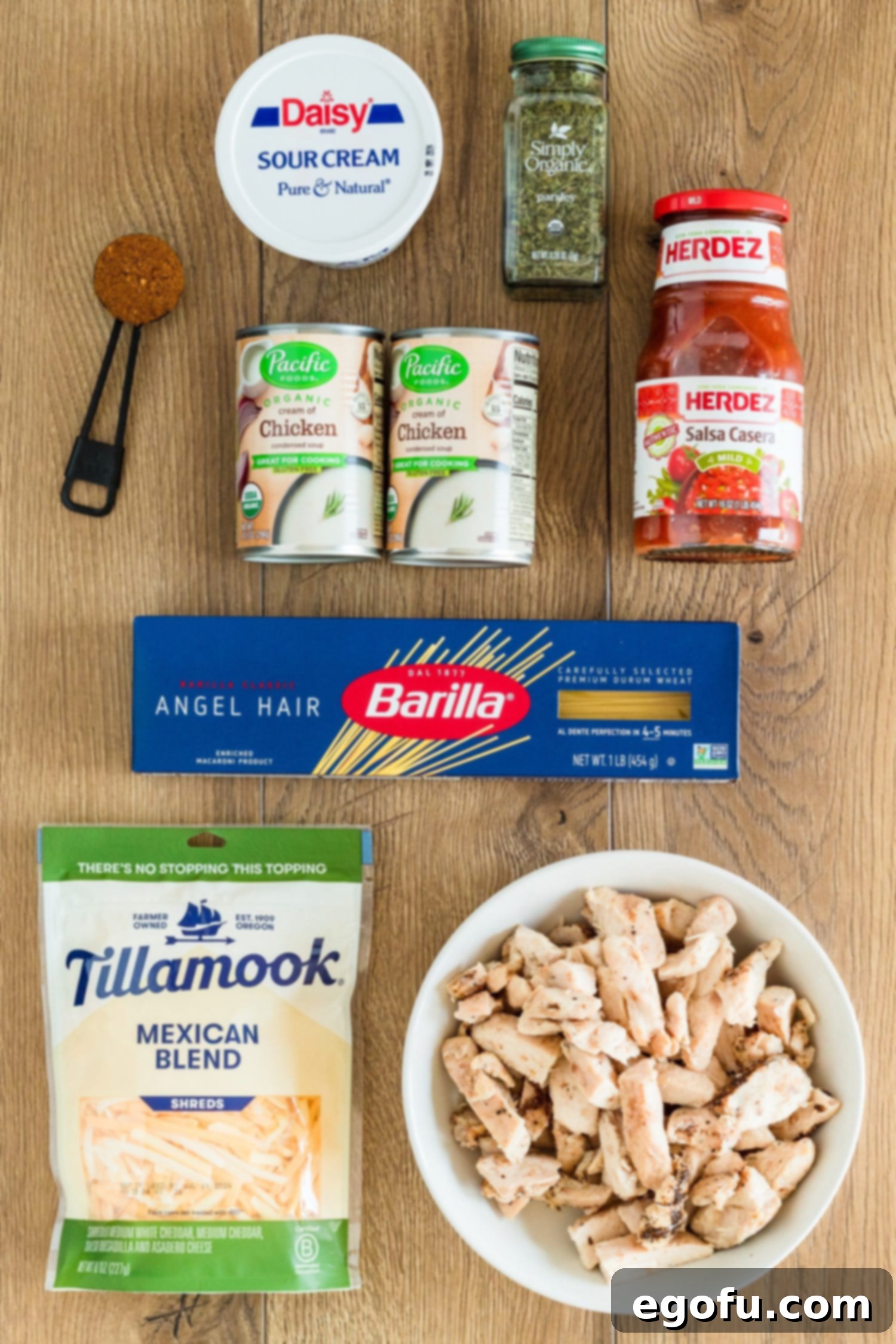 A vibrant flat lay of key ingredients for Chicken Spaghetti: sour cream, Herdez Salsa casera, cream of chicken soup, dried parsley, taco seasoning, angel hair pasta, Mexican blend cheese, and cooked chicken.