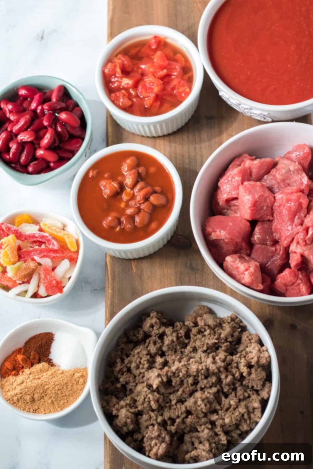 Arrangement of all ingredients for Slow Cooker Ultimate Beef Chili: stew beef, ground beef, canned tomatoes with green chiles, tomato sauce, dark red kidney beans, chili beans, frozen mixed peppers and onions, taco seasoning, chili powder, paprika, and granulated sugar.