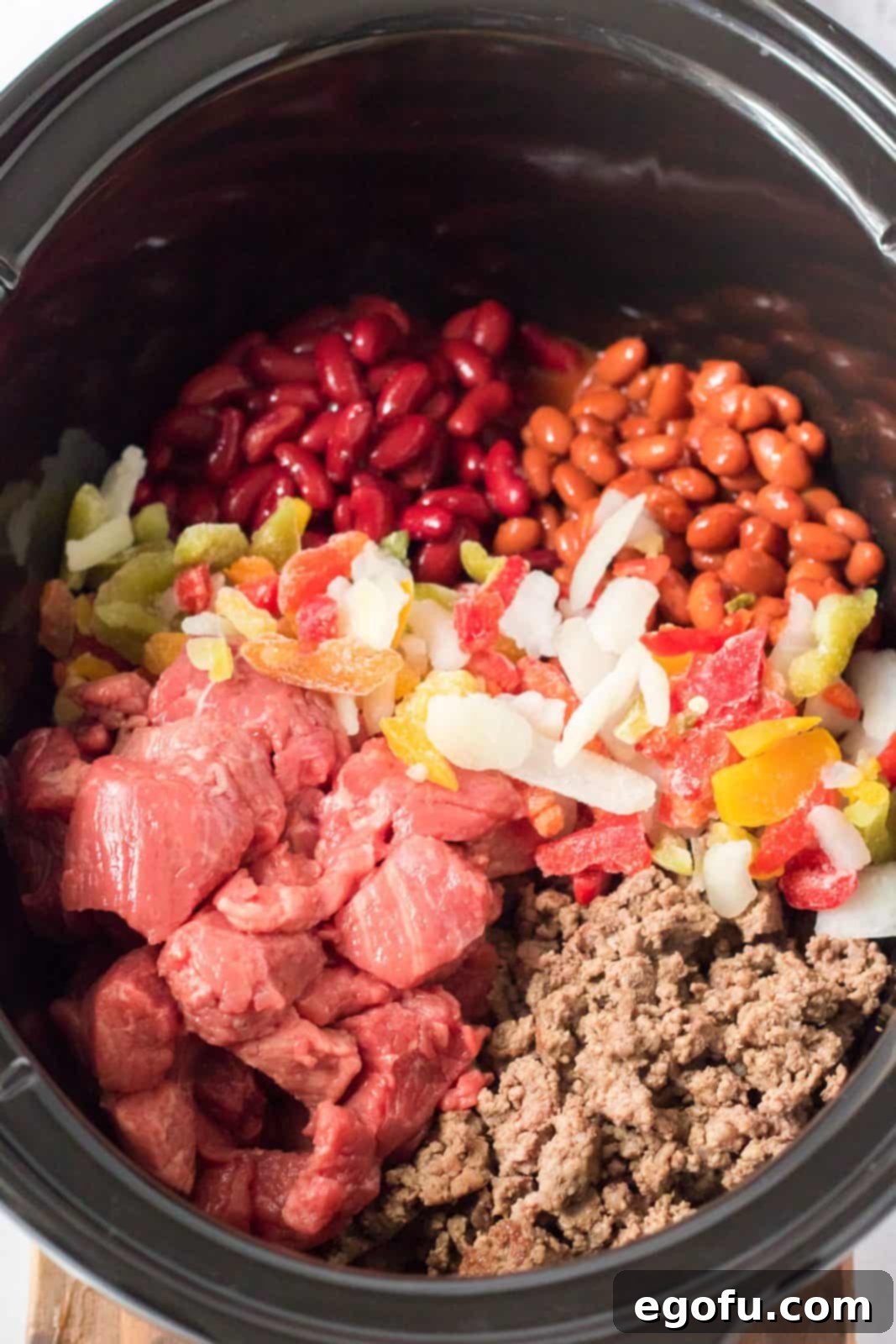 All ingredients, including browned ground beef, stew beef, canned goods, and spices, are combined in a slow cooker, ready for cooking.