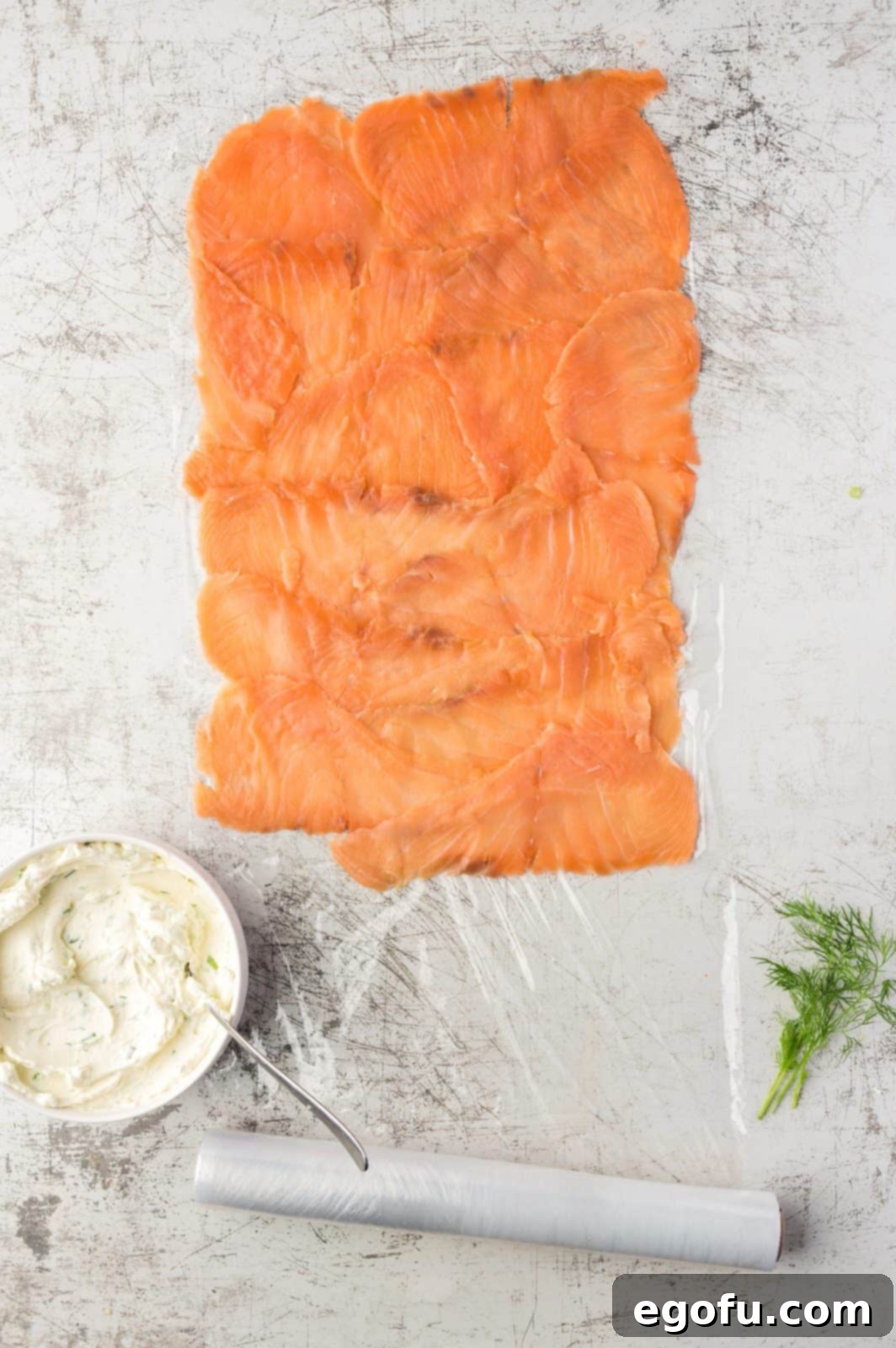 Thinly sliced smoked salmon layered neatly and slightly overlapping on a sheet of plastic wrap, creating a uniform base.