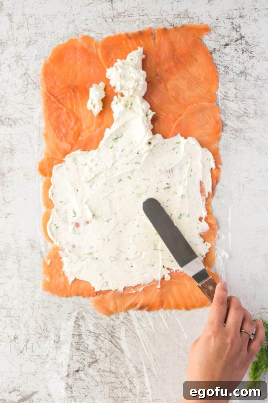 A hand gently spreading the creamy white filling evenly over the laid-out smoked salmon, leaving a clear margin at one end.