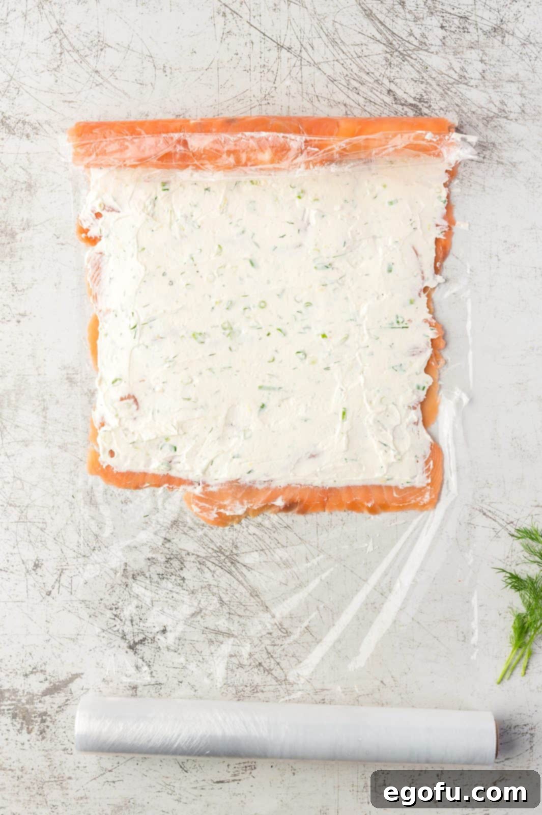 The process of gently rolling up the salmon and its filling into a tight log, guided by the plastic wrap underneath.