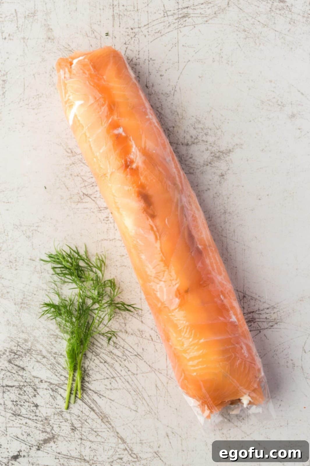 The fully rolled salmon log, tightly wrapped in clear plastic film, resting before chilling to set its shape and flavors.