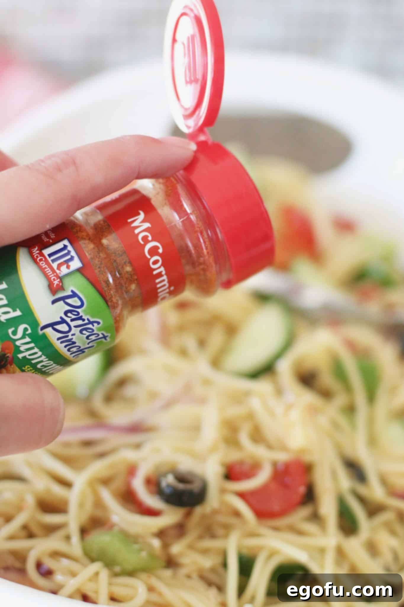 McCormick Salad Supreme Seasoning being generously shaken over a freshly prepared bowl of spaghetti salad, enhancing its flavor profile.
