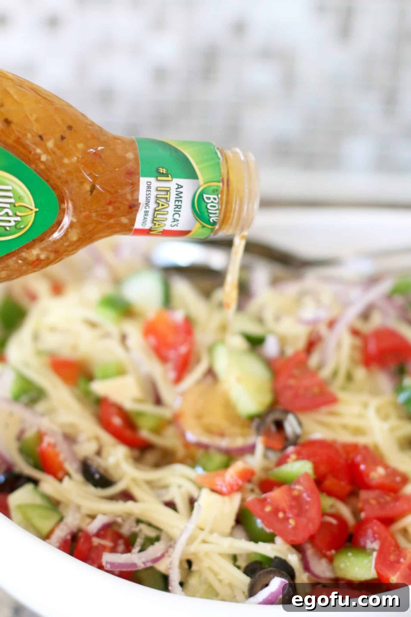 Wish-Bone Zesty Italian Dressing being poured generously over a bowl of vibrant spaghetti noodle salad, ready for mixing.